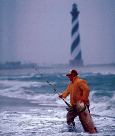 Photograph of a man fishing off the coast of North Carolina; Cape Hatteras lighthouse in the background.